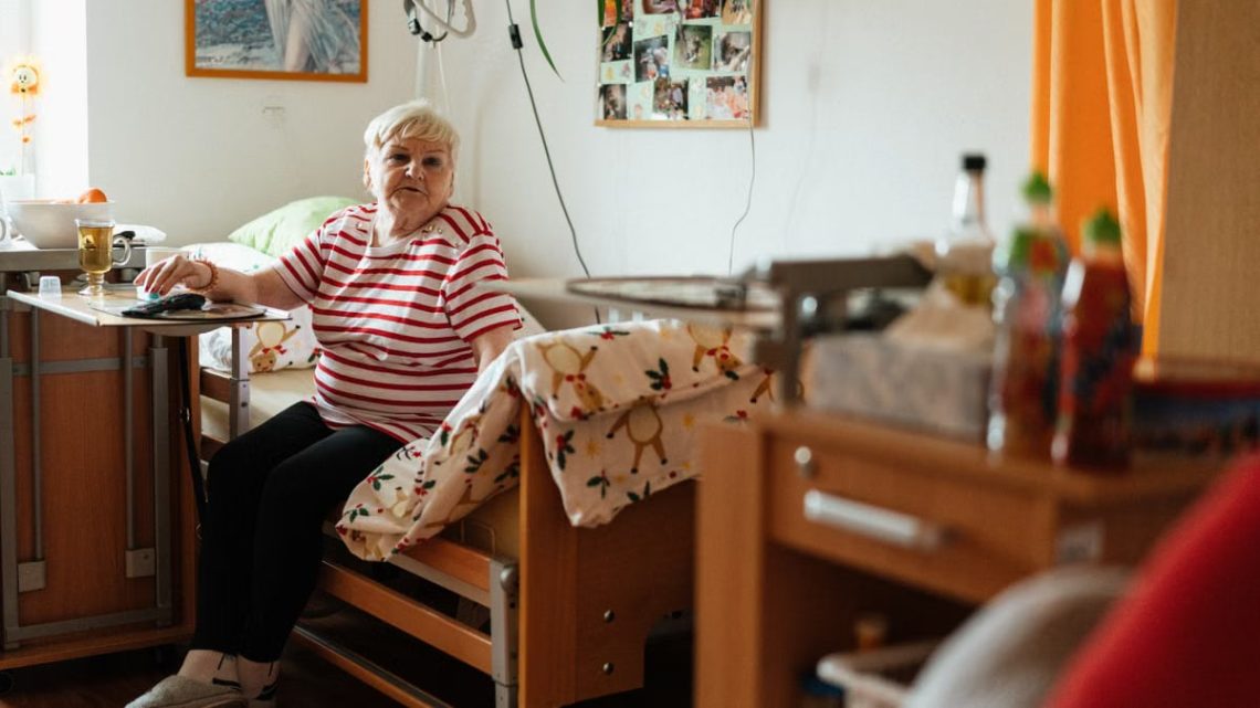 Elderly woman in striped shirt sits comfortably in her retirement home bedroom, surrounded by personal items.